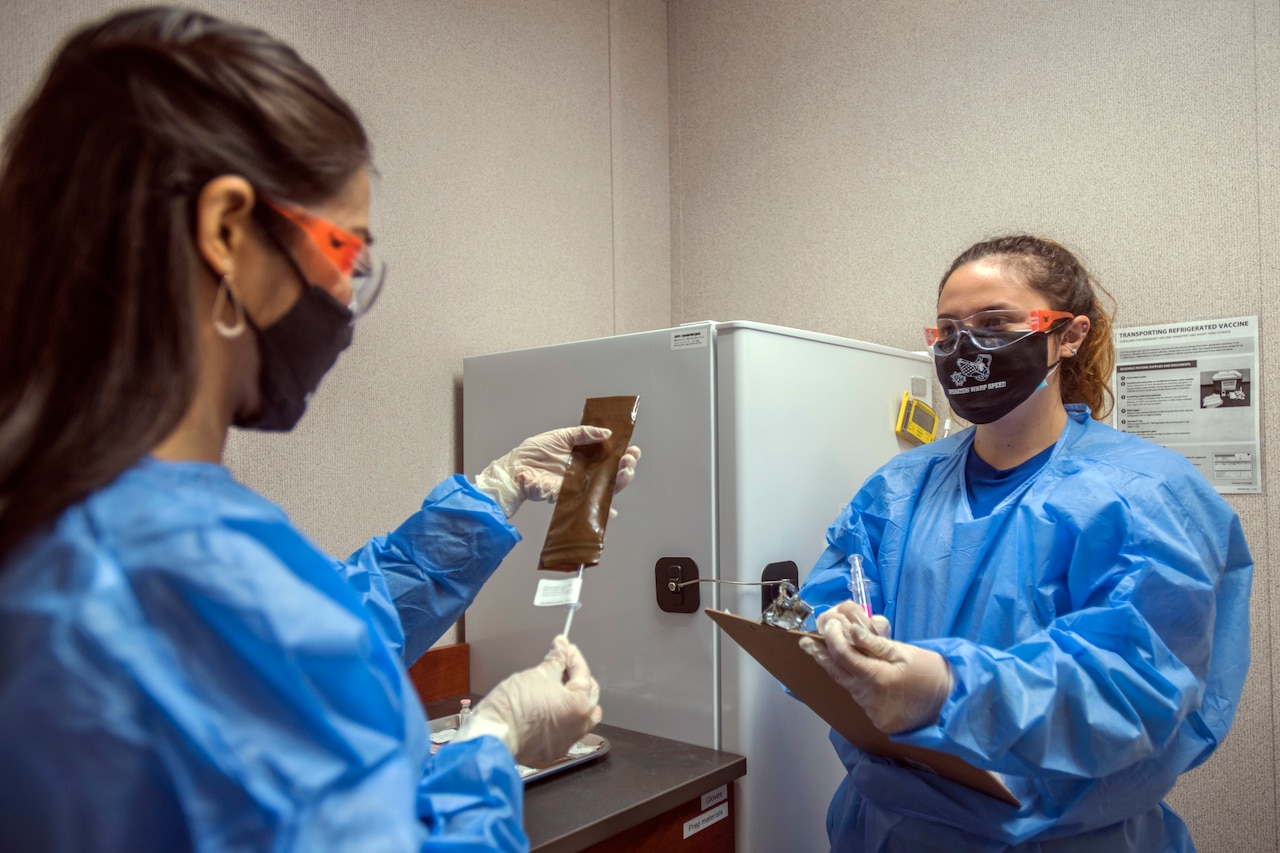 A woman wearing personal protective equipment documents and verifies if a patient has received a vaccine or placebo. Another woman holds a clipboard.