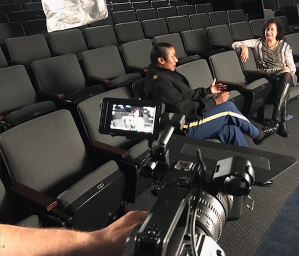 Two women sit in auditorium chairs talking with a video camera in the foreground.