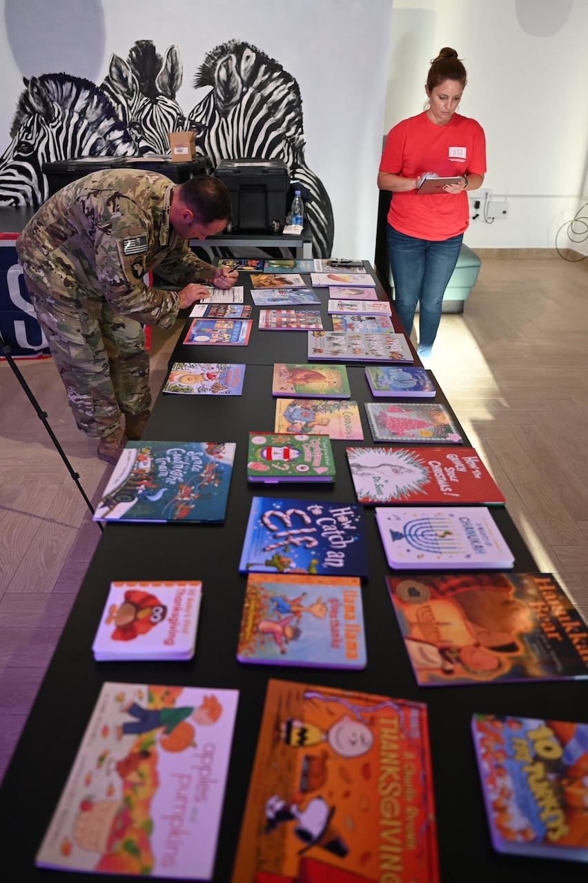 A military man writes a note near children's books as a woman works on an ipad.