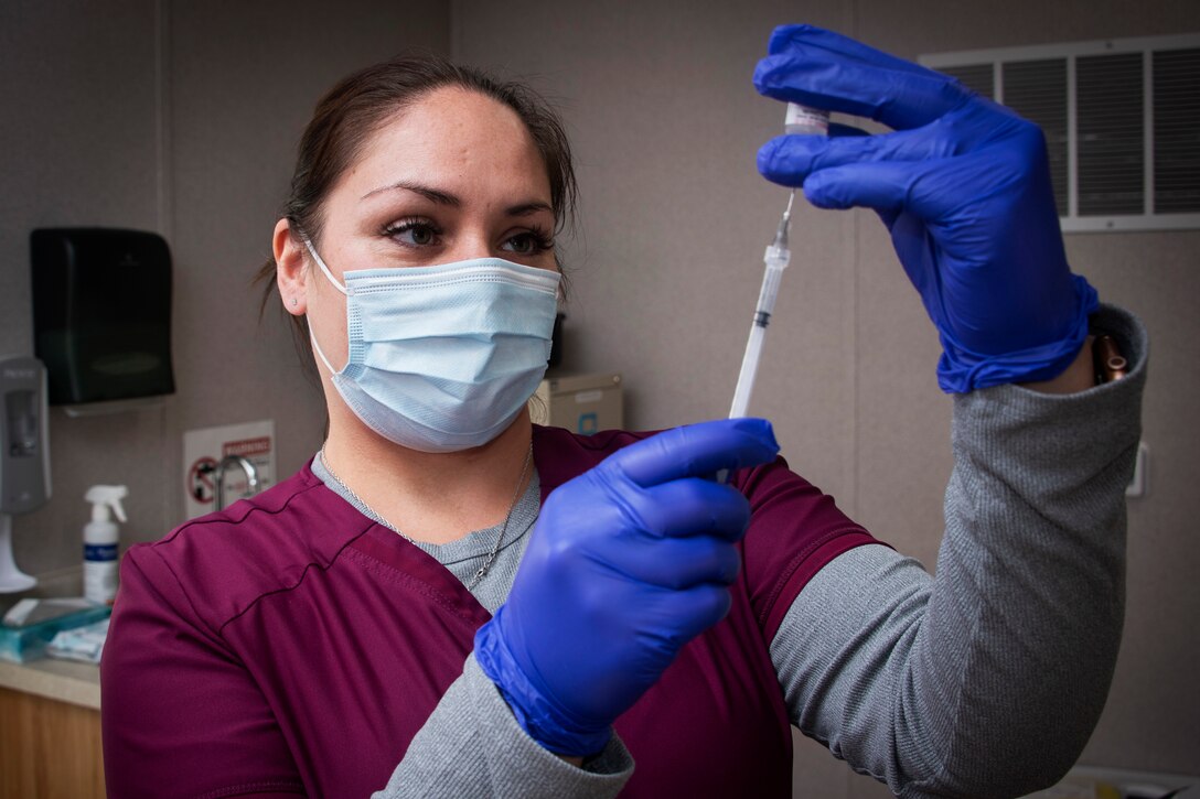 A pharmacy technician wearing a face mask and gloves prepares a syringe for a patient.