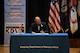 Two men signing contractual agreement on a stage.