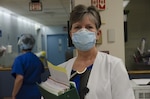 A nurse poses inside a nursing home facility while wearing a white lab coat and a face mask. She is holding a large binder full of folders and papers.