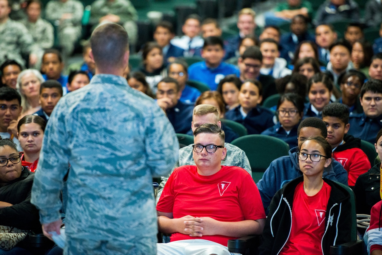 Young students sit in rows in an auditorium and listen to a military officer speak.