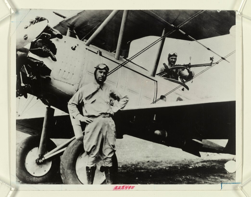 An aviator stands beside an old propeller plane as another man works in the cockpit.