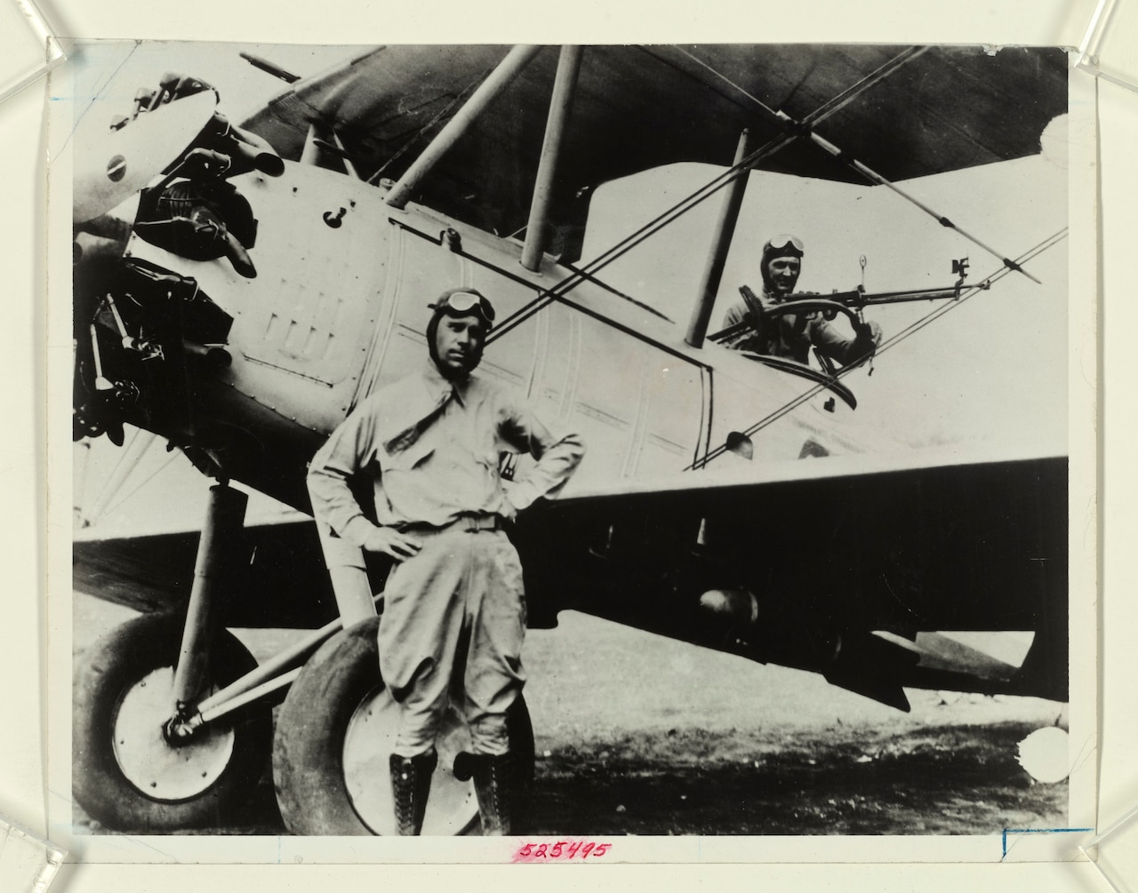 An aviator stands beside an old propeller plane as another man works in the cockpit.