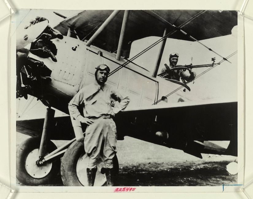 An aviator stands beside an old propeller plane as another man works in the cockpit.