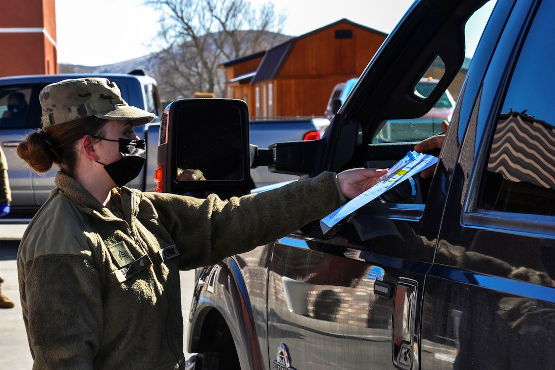 A soldier wearing a face mask hands a piece of paper to someone through the driver's side window of a vehicle.