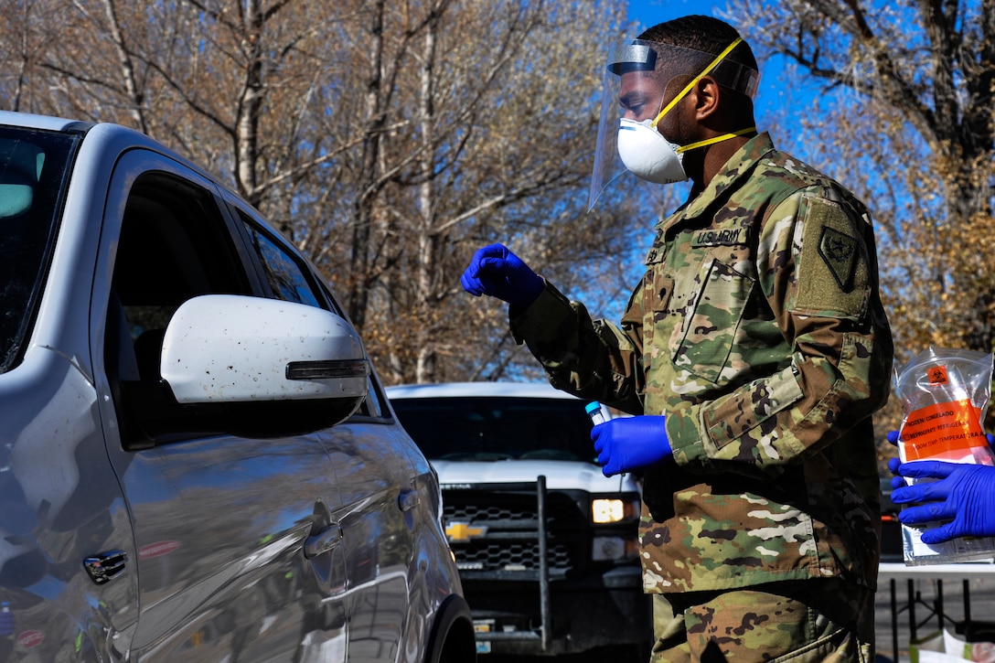 A soldier wearing a face mask and gloves assist with giving a patient a nasal swab test in their vehicle