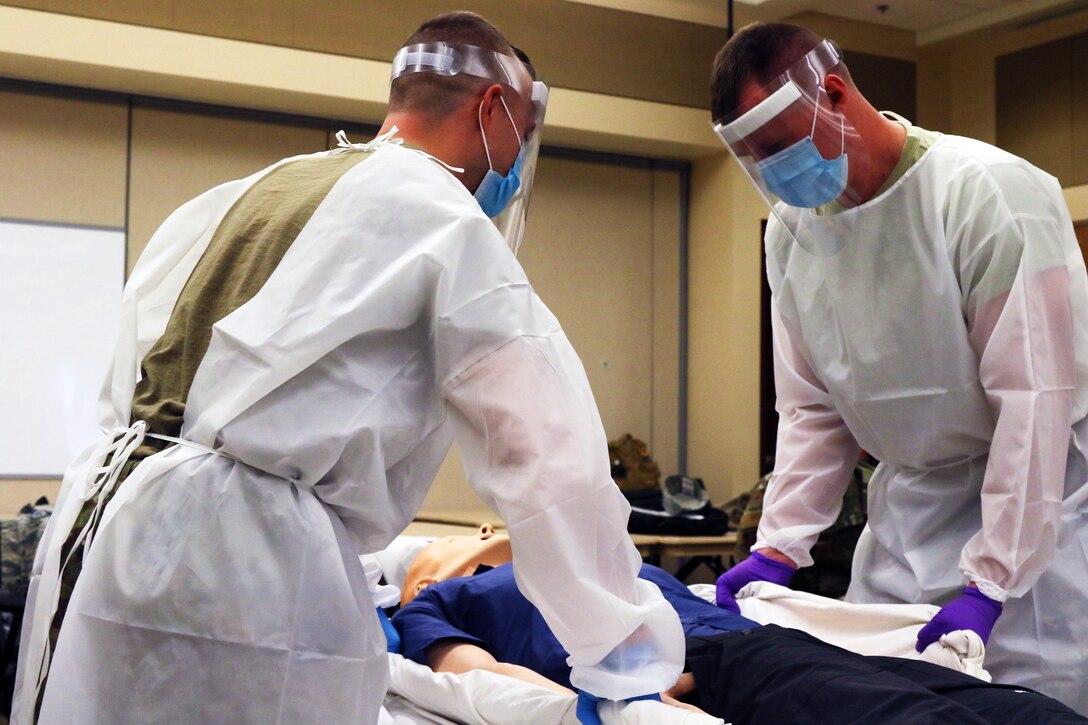 Two soldiers wearing personal protective equipment stand on either side of a mannequin on a gurney.