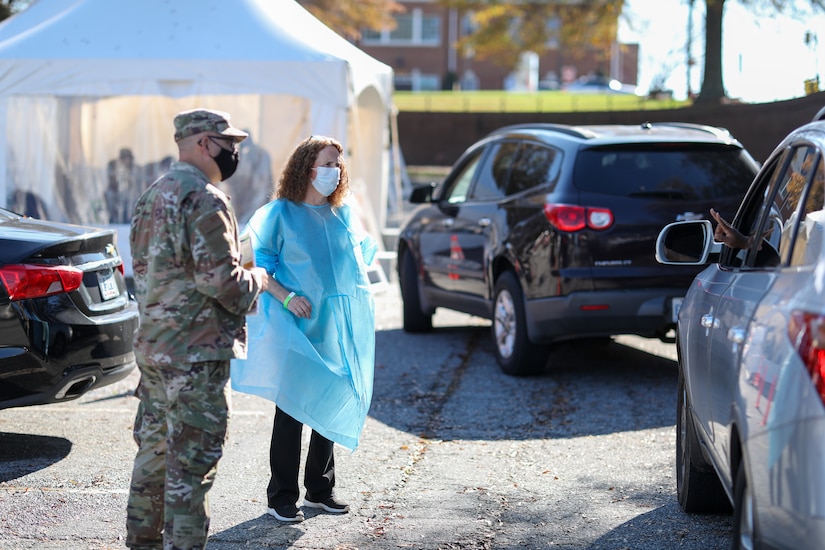 A civilian woman and a soldier talk to patients at a drive-thru.