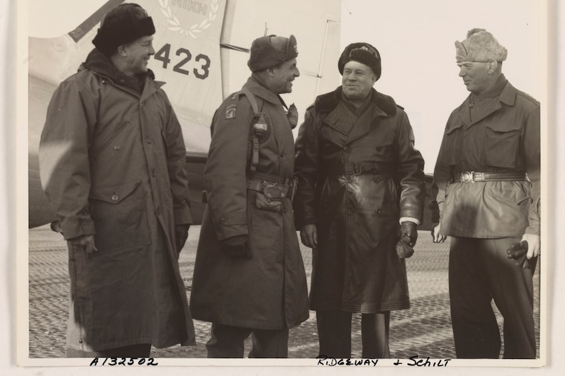 Four men in winter garb stand beside the tail of an airplane.
