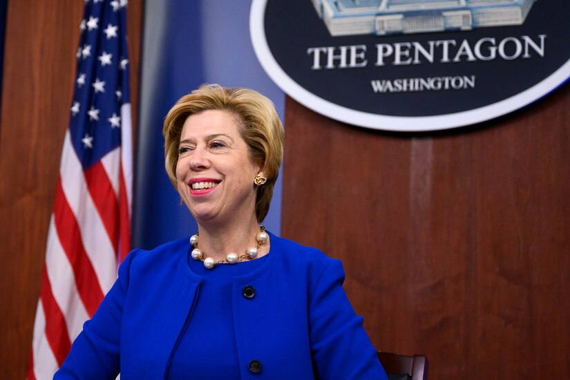 A woman in a blue dress stands in front of a U.S. flag and a sign that reads "The Pentagon - Washington."