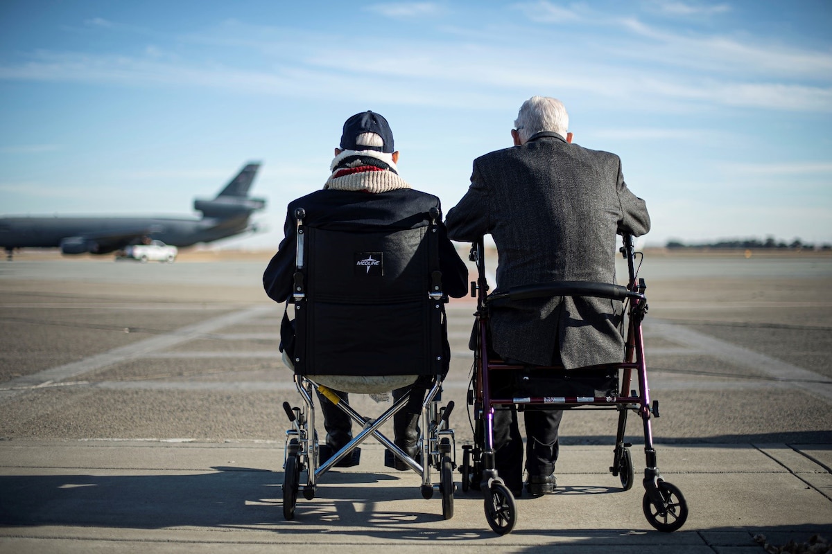 Two veterans sit in chairs on a flightline and watch an aircraft taxi.