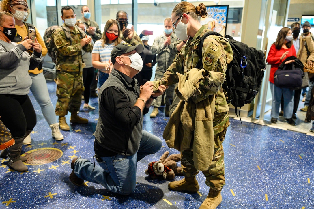 A man kneels in front of a service member as crowd watches.