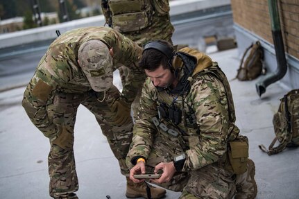 A service member crouches on the ground to look at an instrument in his hand as another service member leans down to observe.