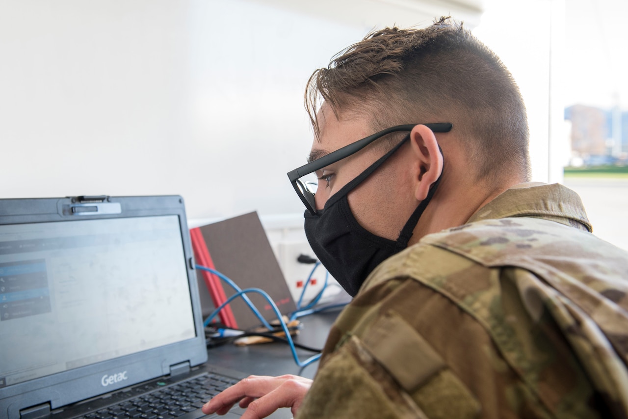 An airman wearing a face mask works on a laptop.