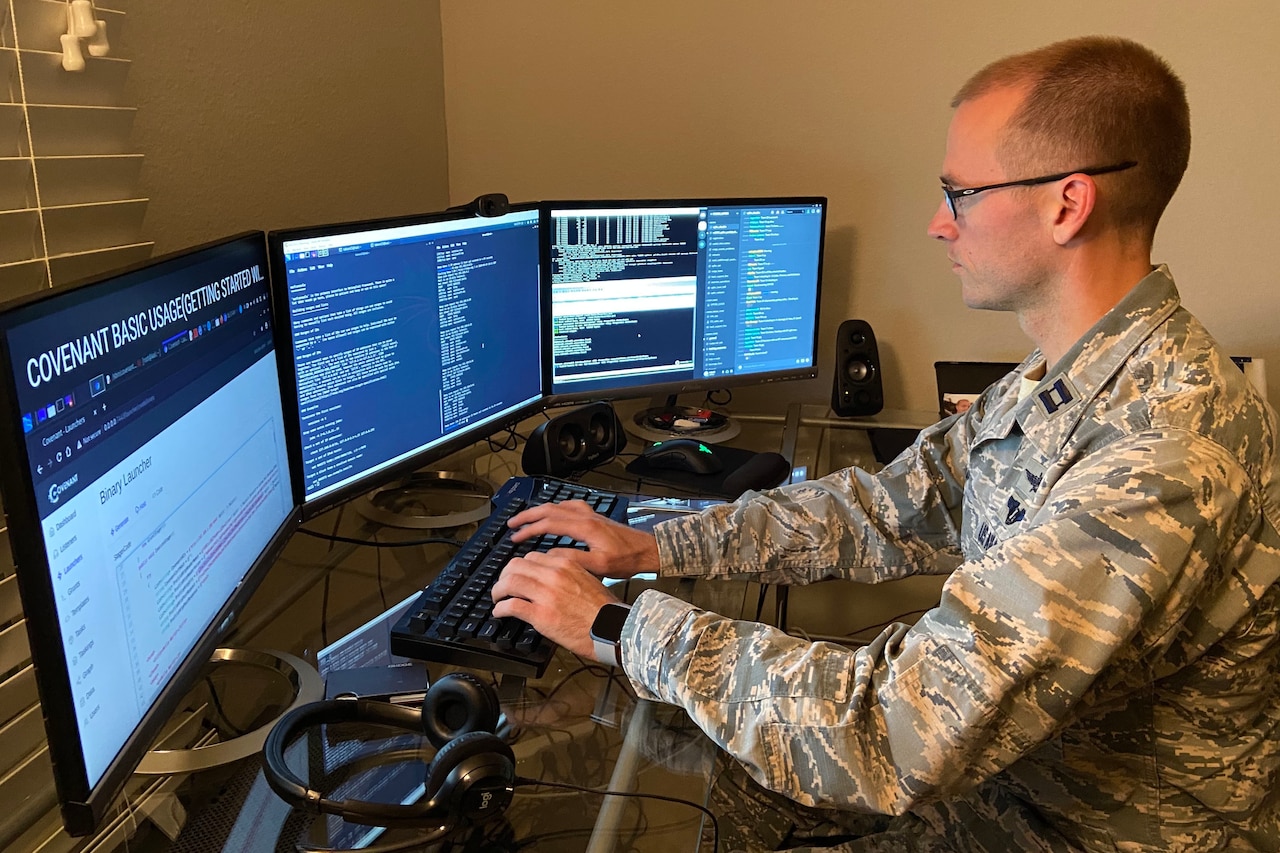 A man in a military uniform types on a keyboard and looks at three monitors.