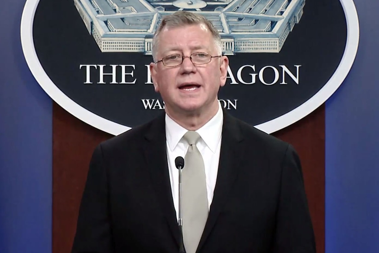 A man stands at a lectern and speaks into a microphone. A sign indicating that he is at the Pentagon hangs on the wall behind him.