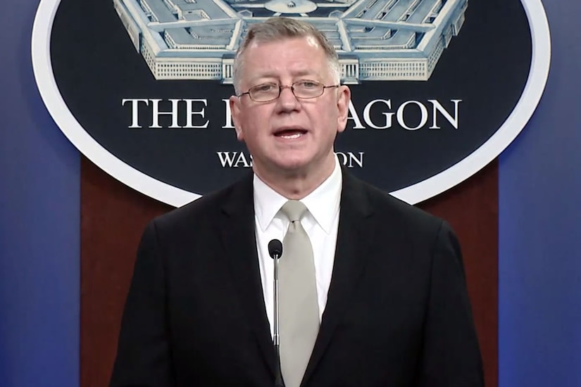 A man stands at a lectern and speaks into a microphone. A sign indicating that he is at the Pentagon hangs on the wall behind him.