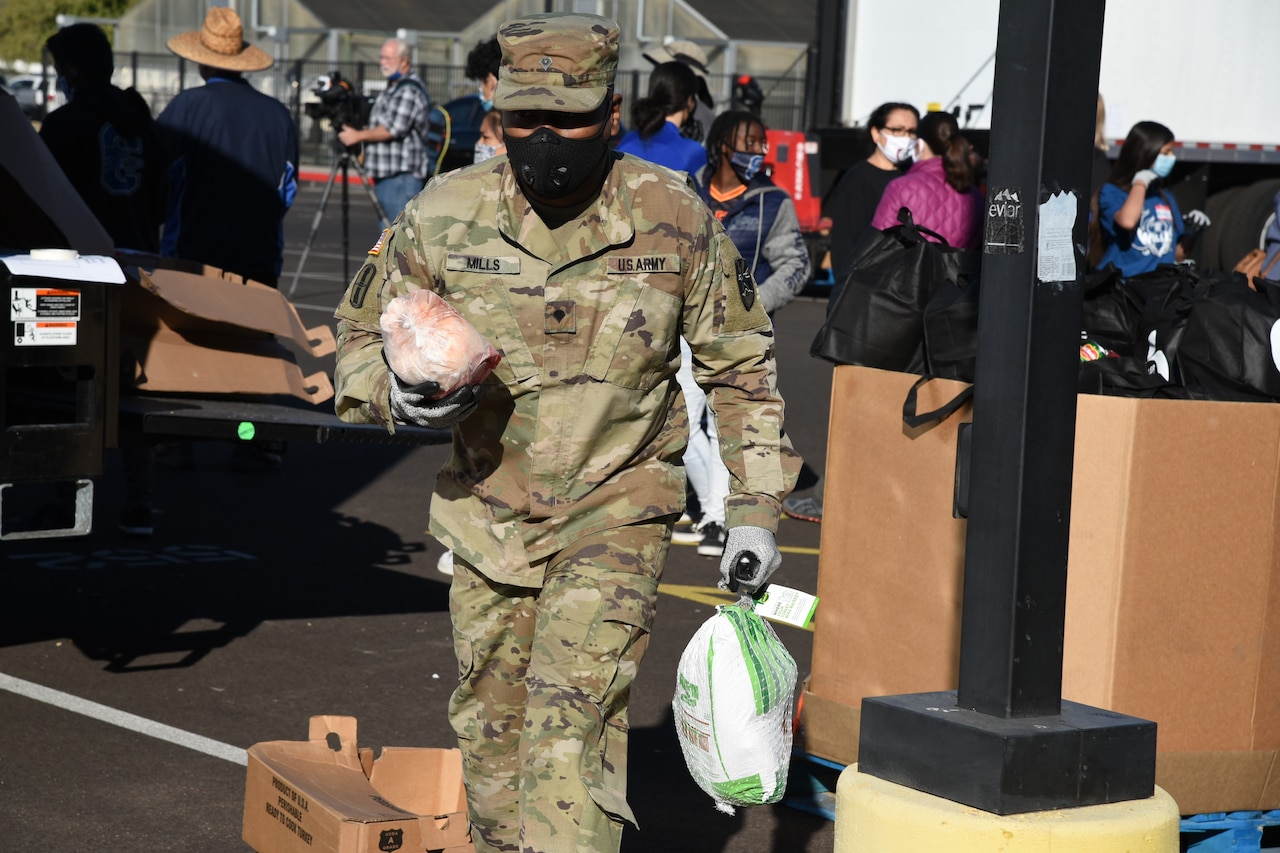 A soldier wearing a face mask carries two frozen turkeys across a parking lot.