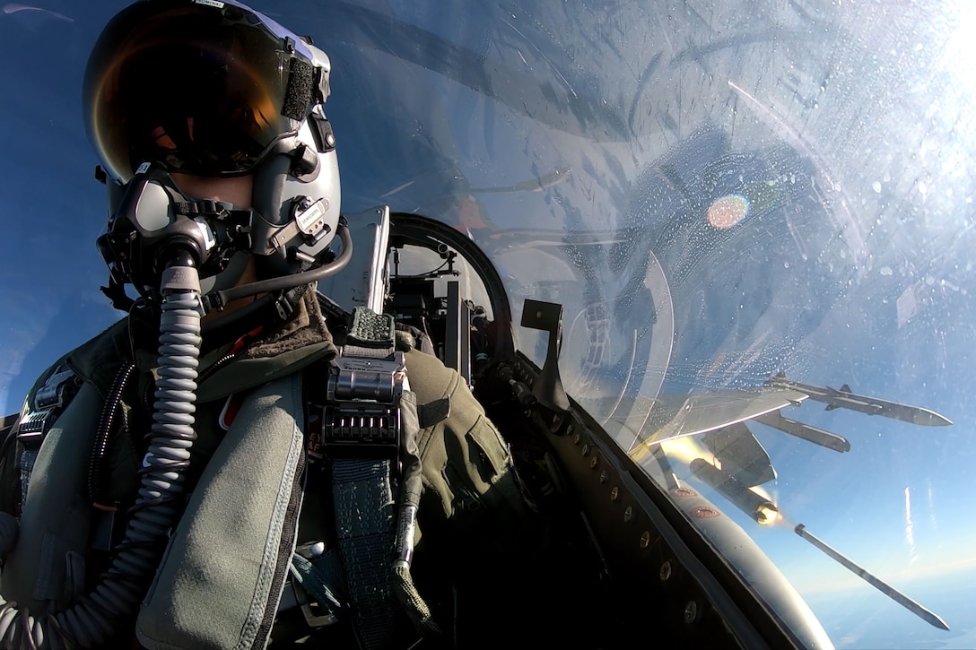 A military pilot wearing an oxygen mask sits in an aircraft cockpit while firing a rocket from his plane.