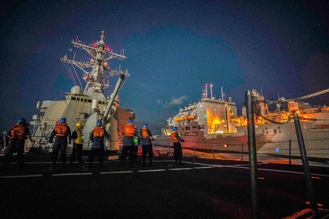 Service members in life vests stand on a ship while observing another one.
