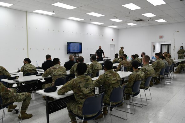 Photo shows students sitting at long tables being taught by a person standing in front of the room.