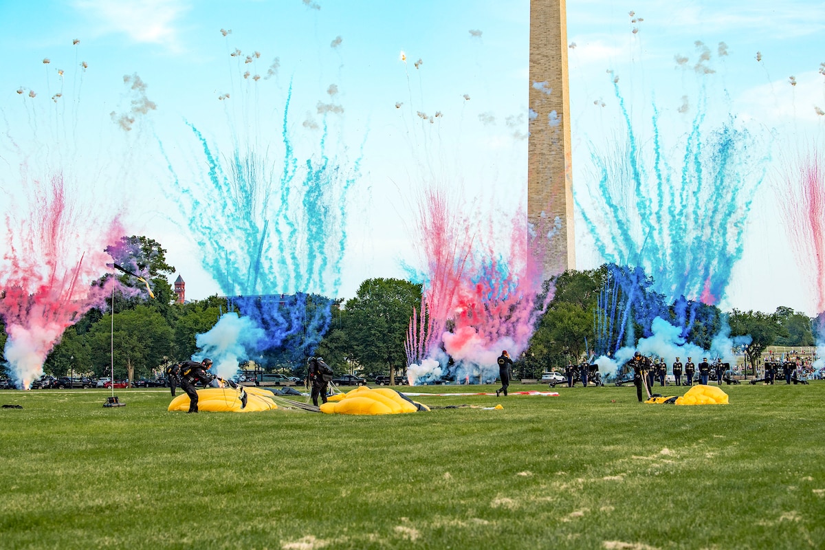 Blue and pink fireworks erupt as soldiers gather their parachutes on grounds in front of the Washington Monument.