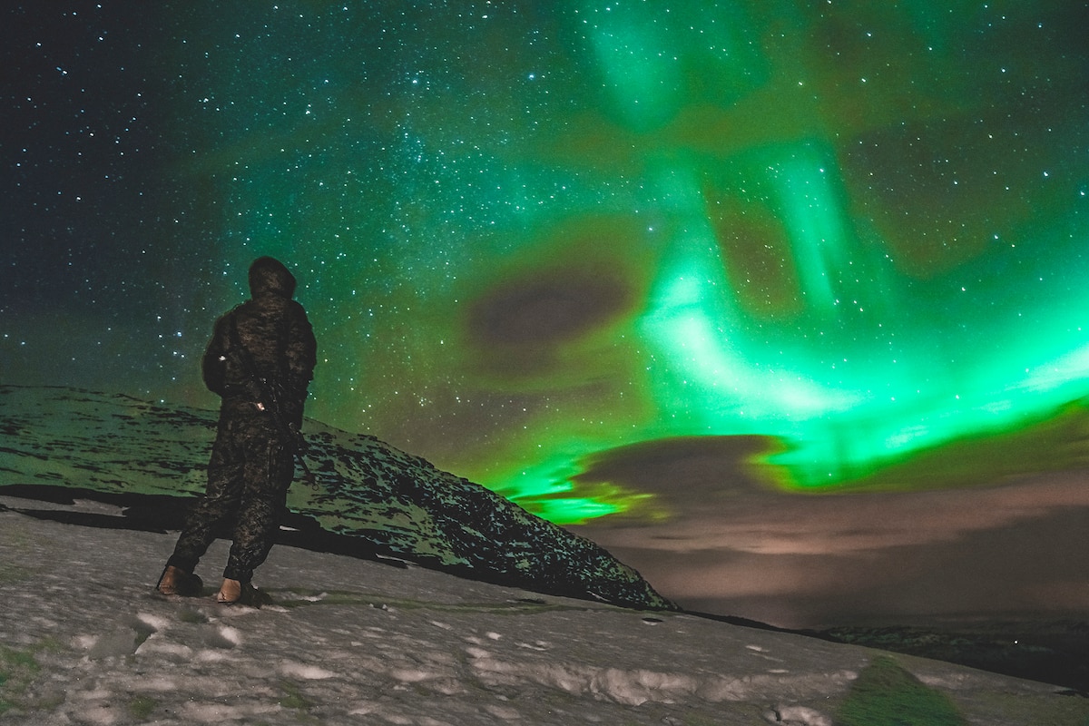 A Marine stands in snow and looks toward the night sky, illuminated in green by the Northern Lights.