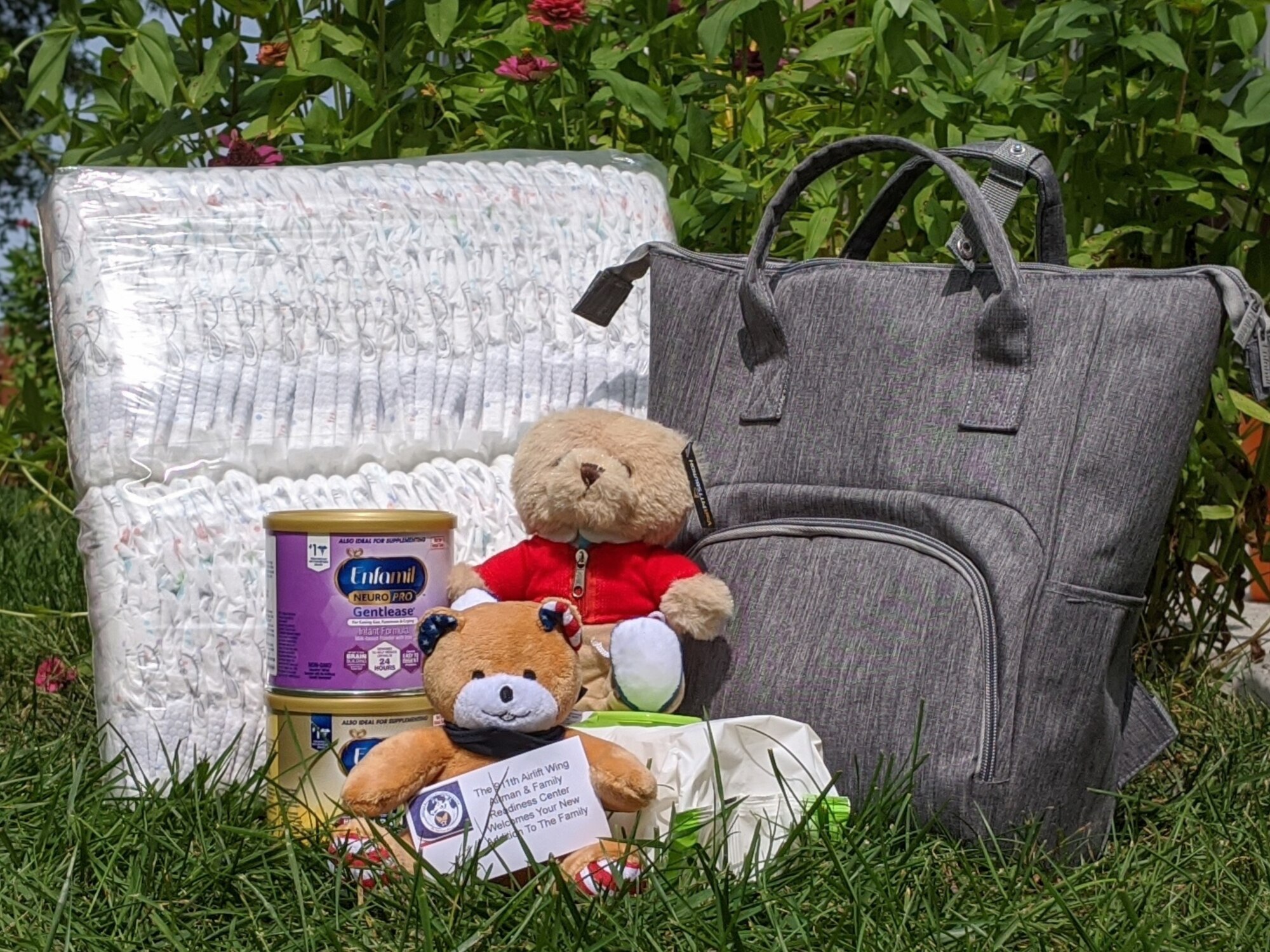 A blanket, diaper bag, two teddy bears, two containers of baby formula, and a package of baby wipes arranged on a grassy lawn with a flowering shrub in the background.