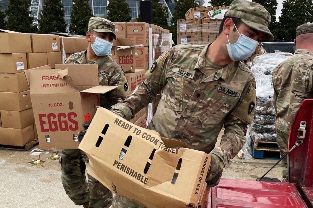 Two guardsmen wearing face masks and gloves carry boxes of food at a distribution point.