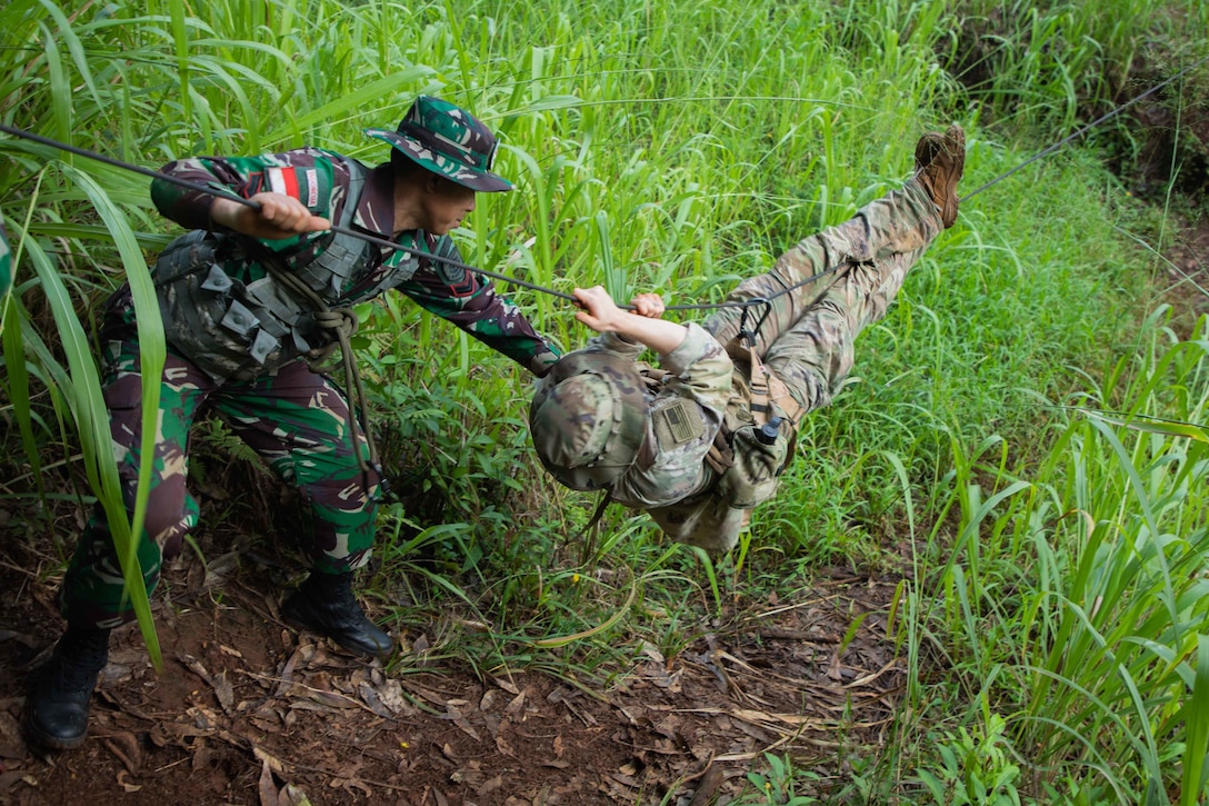 A soldier clings to a rope with his hands and feet while another soldier  watches closely.