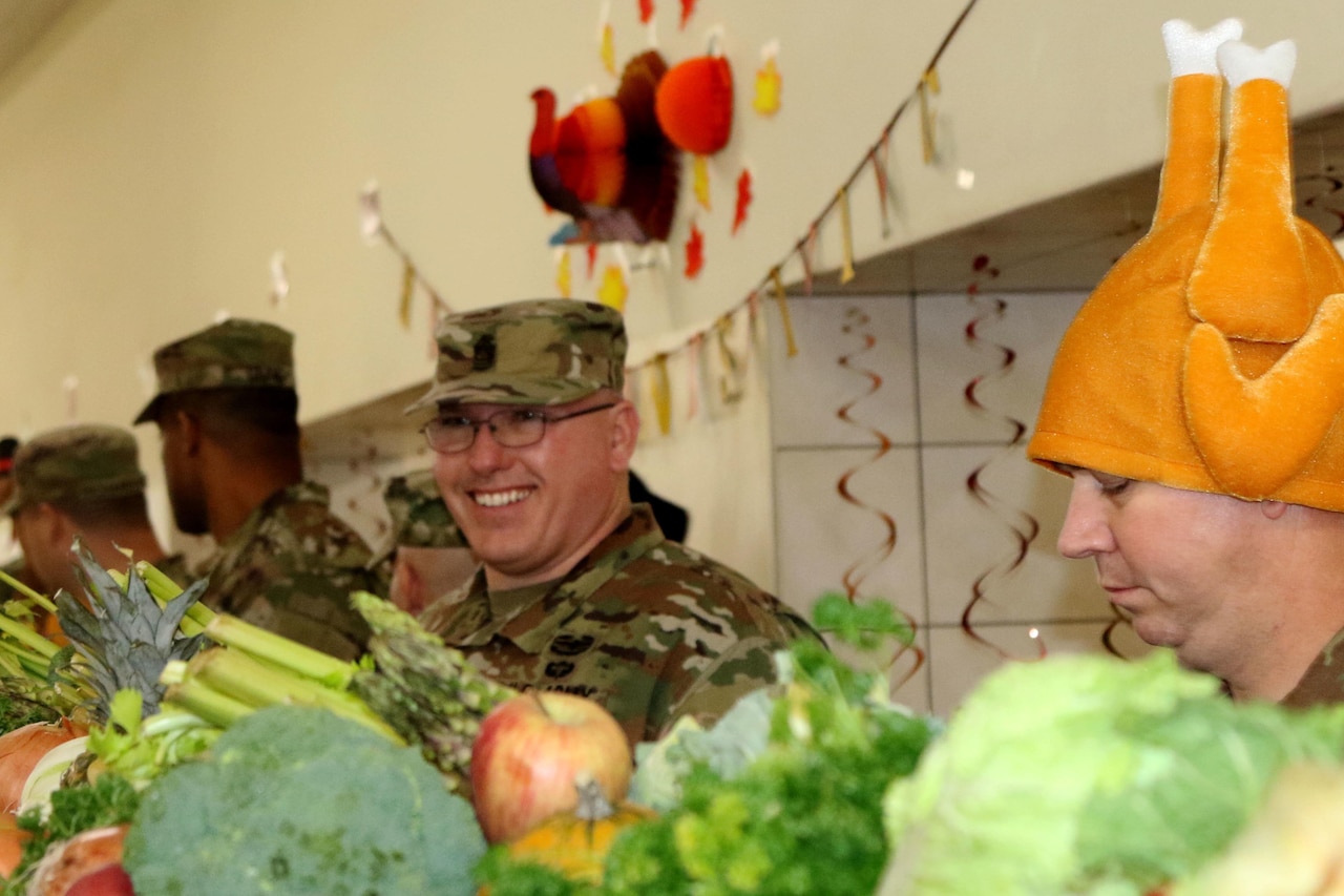 A soldier smiles behind a tray of vegetables. Next to him is a man wearing a turkey-leg hat.