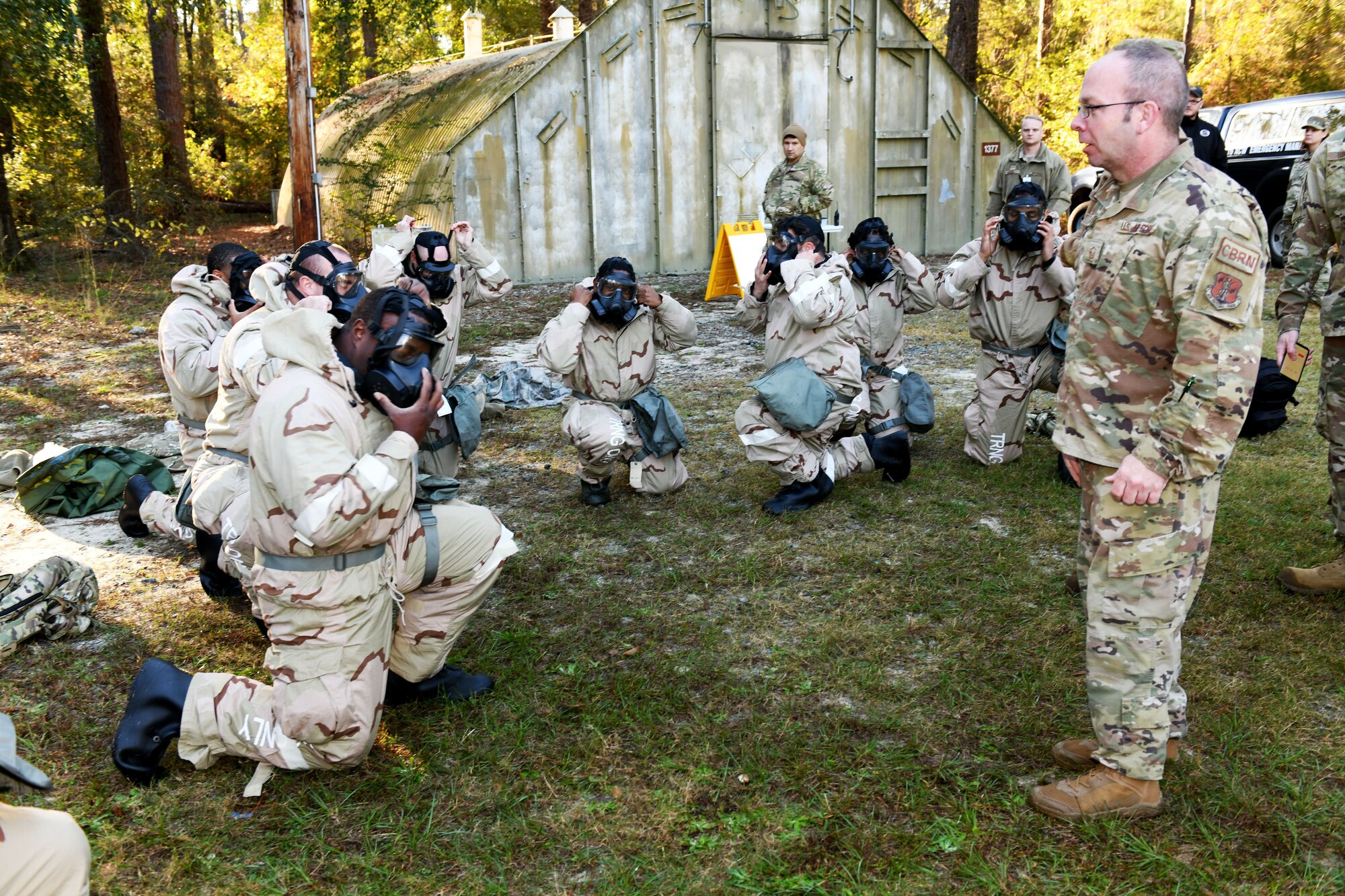 Photo shows Airmen kneeling while placing chemical gear on and an instructor watching.