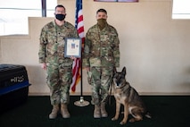 Two uniformed men stand with a certificate in hand, with the military working dog sitting by their side.