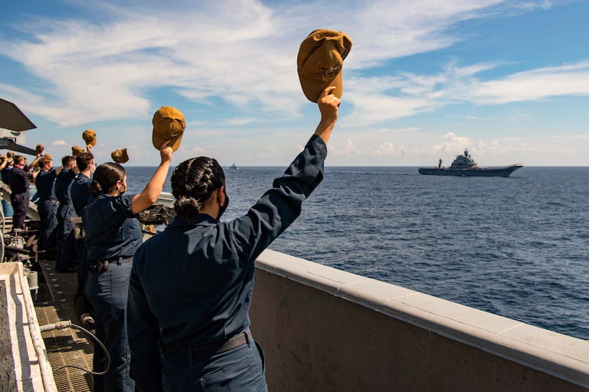 Sailors standing on a ship wave their hats at a ship sailing by.