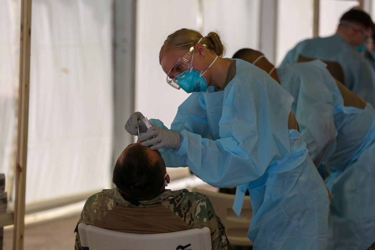 A soldier wearing protective gear leans over a man to conduct a COVID test.