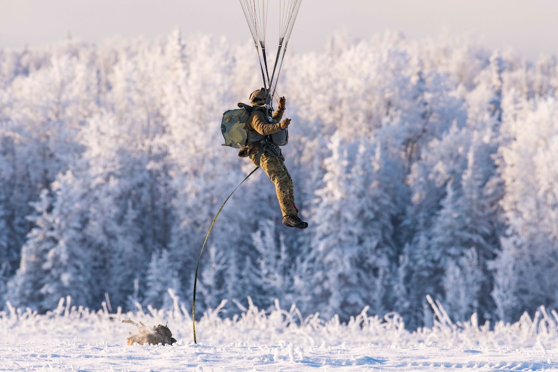 An airman descends from the sky with a parachute onto a snow-covered field with snow-covered trees in the background.