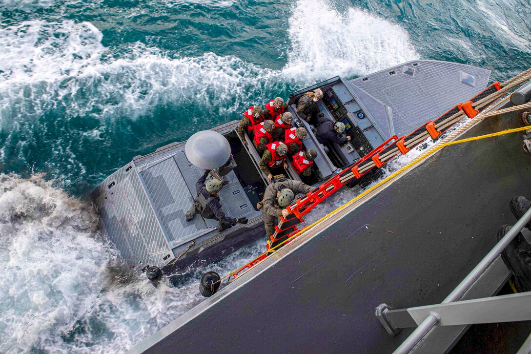 Sailors sit in a boat as another sailor descends a ladder to board it.