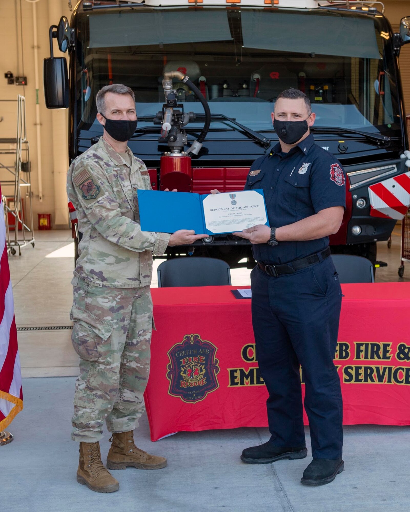 one male colonel in military uniform, stands with a firefighter while holding a certificate while standing in front of a fire truck.