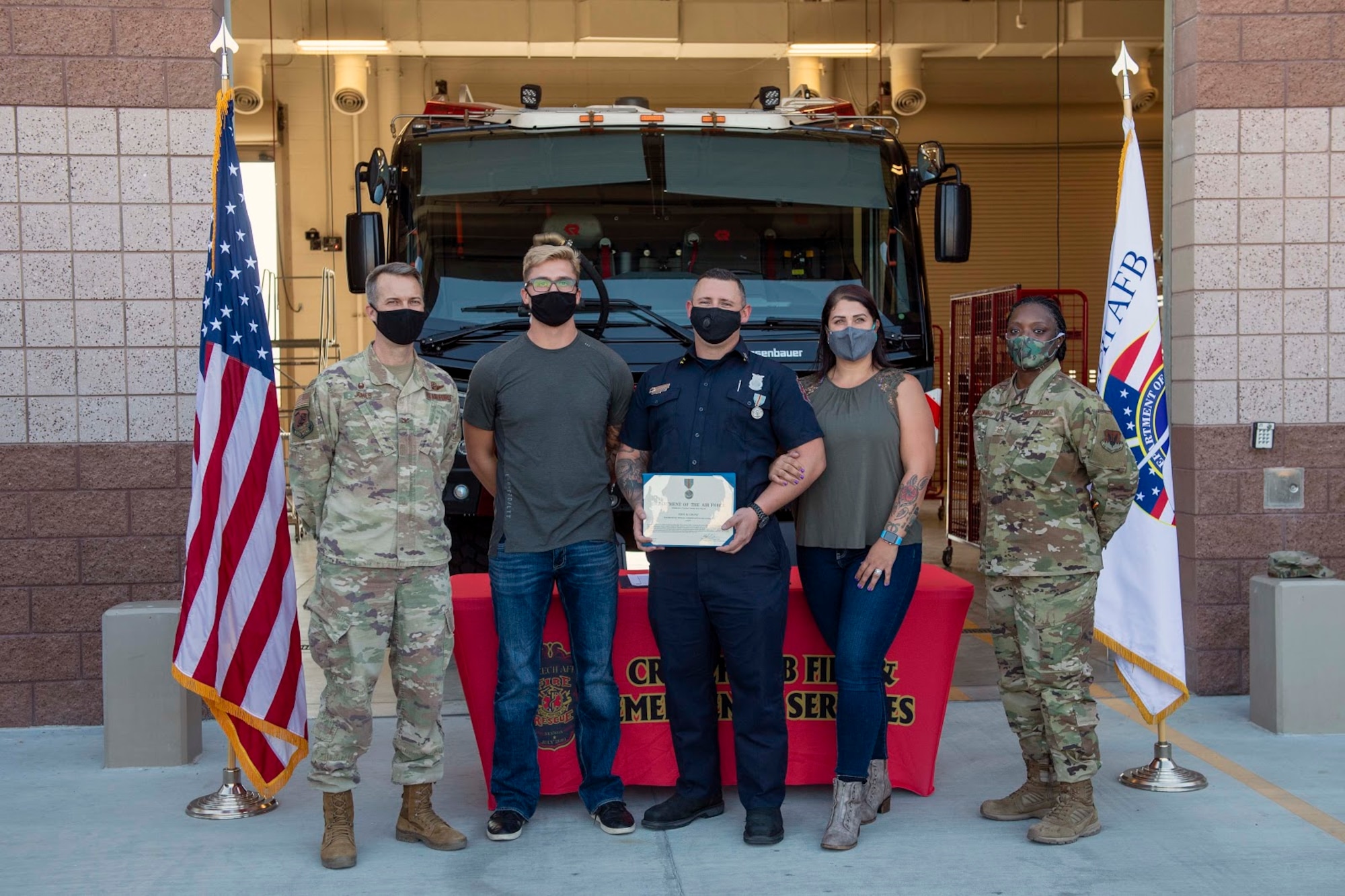 5 adults stand in front of a Fire Truck with the award recipient holding a certificate.