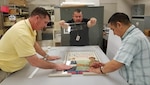 (From left) Museum specialist Chris Goodrow, exhibit specialist Lupe Perez, and archivist Carlos Alvarado work on the finishing touches for featured posters on display in the World War I gallery exhibit at the U.S. Army Medical Department Museum at Joint Base San Antonio-Fort Sam Houston.