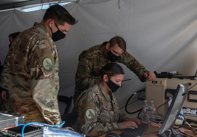 Three airmen study a computer under a tent.