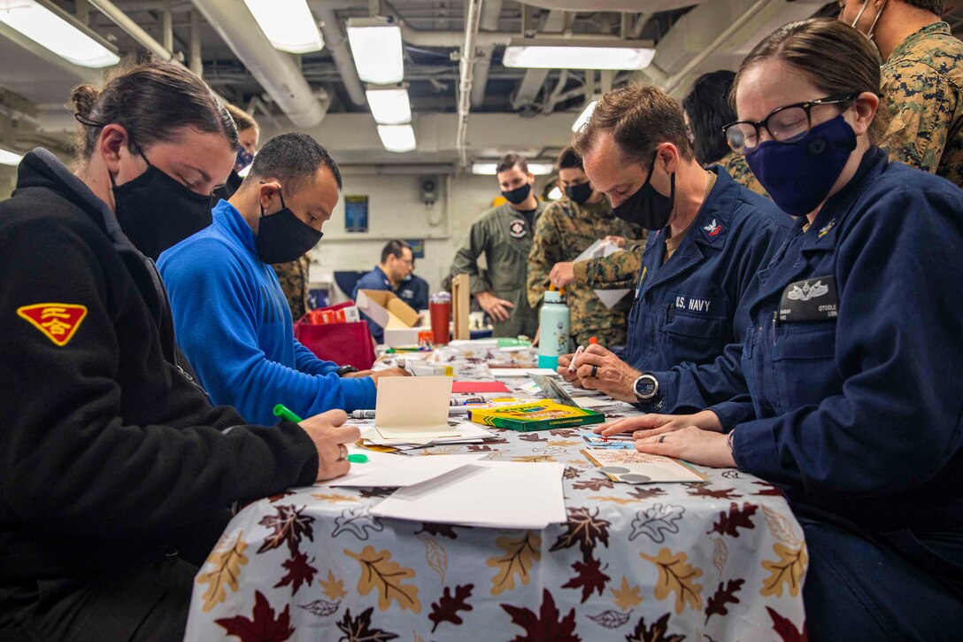Sailors and Marines sit at a table using paper and markers to create holiday cards.