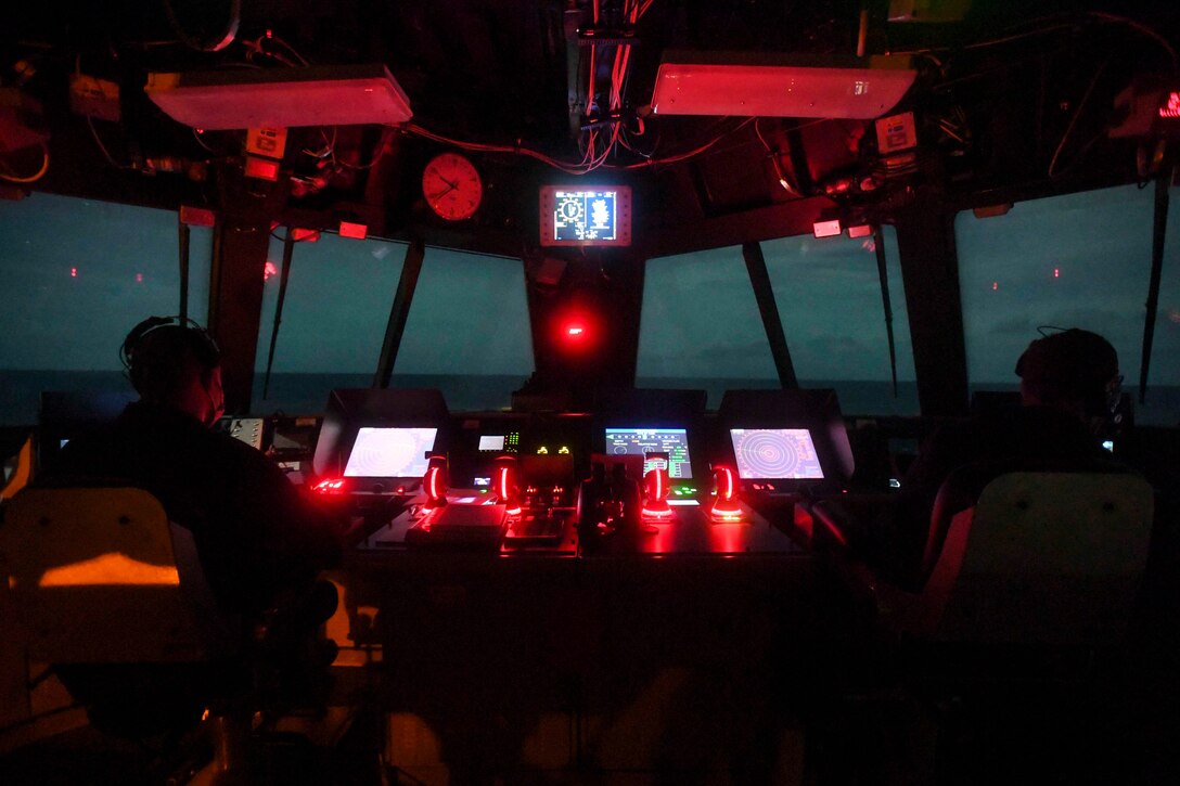 Two sailors sit in the navigation bridge of a ship illuminated by monitors and red light at night.