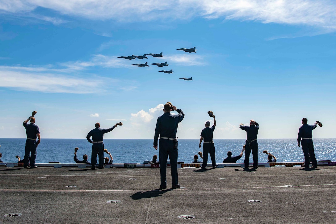 Sailors standing on the deck of a ship wave at eight aircraft flying above.