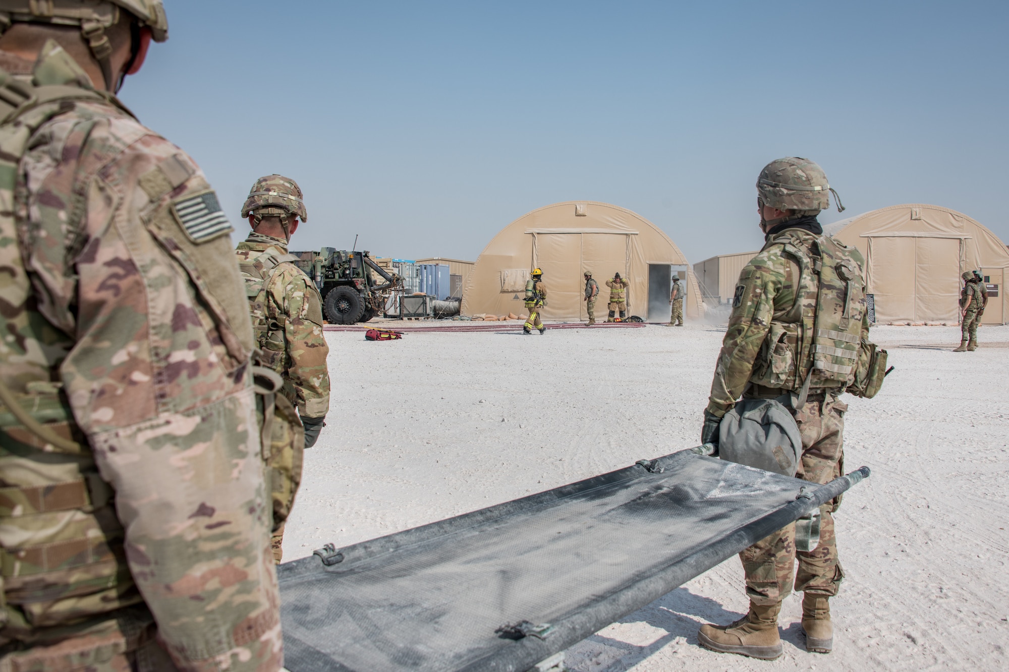 Soldiers wait with a stretcher for firefighters in the background to clear a building full of simulated smoke