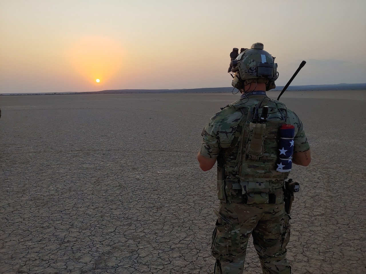 A man is seen from behind staring toward the sunset; in front of him, the ground is dry and cracked.