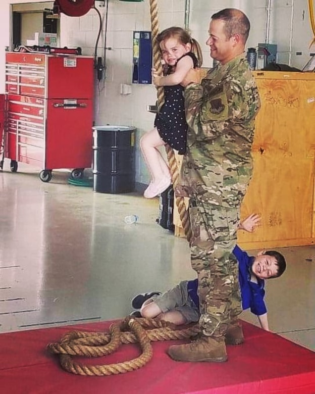 An airman lifts a small girl, who is grasping a large rope that dangles from above. A small boy waves to the camera from beside the man's feet.