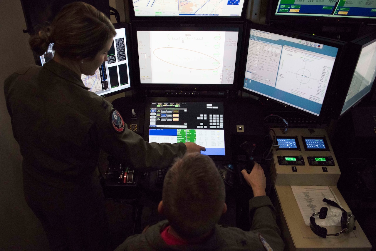 A man in a military uniform sits inside a simulated aircraft cockpit. A woman in a military uniform stands beside him.